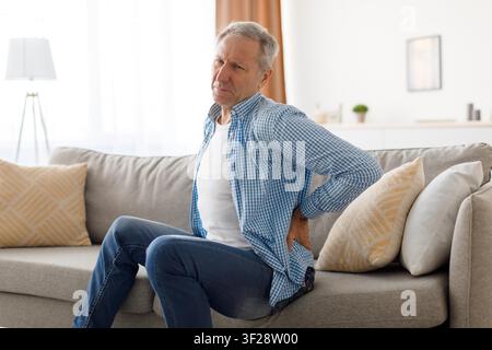 Elderly Caucasian Man Experiencing Back Pain While Sitting On Couch At Home, Healthcare Concept Stock Photo