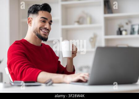 Smiling Young Man With Beard Drinking Coffee While Using Laptop At Home, Business Concept Stock Photo