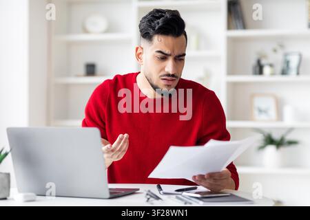 Concerned Young Arab Man Reviewing Documents At Desk, Office Setting, Business Concept Stock Photo