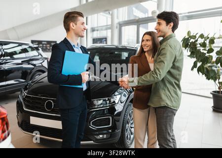 Happy Young Caucasian Couple Shaking Hands With Car Salesman In Dealership, Business Concept Stock Photo