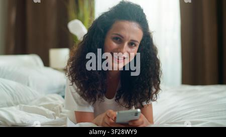 Caucasian relaxed calm woman using mobile phone app smiling in bedroom at home happy girl female browsing smartphone social media chatting smile Stock Photo