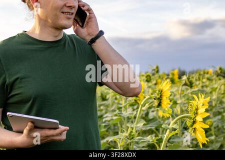 Close up of middle aged man talks on smartphone while holding tablet during crop inspection in blooming sunflower field. Concept of agricultural commu Stock Photo