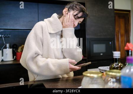 Upset woman in kitchen getting bad news on her mobile phone, feeling sad and anxious Stock Photo