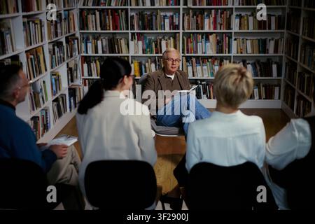 Senior Caucasian man leading group discussion in library, holding book and guiding adult participants in learning session. Useful for education, coaching, book club, community workshop Stock Photo