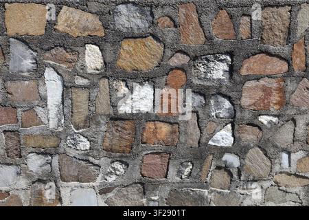 Natural stone wall with flat sandstone, porphyry, calcareous rock, and white quartzite assembled in dark cement mortar.  Background and texture. Stock Photo