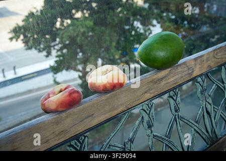 Flat Donut Peaches and Green Mango Resting on Wooden Balcony Railing Above City Street Stock Photo