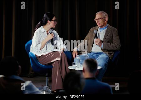 Young adult woman interviewing senior Caucasian man on stage, holding microphones and discussing expertise before audience. Ideal for business events, leadership talks, media coverage Stock Photo