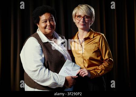 Portrait of middle aged Black woman and Caucasian woman holding book and facing camera, representing partnership and leadership. Useful for diversity, publishing, mentoring campaigns Stock Photo