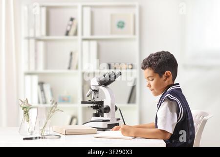 Schoolboy sitting at a desk with a microscope and plants and writing in a notebook Stock Photo