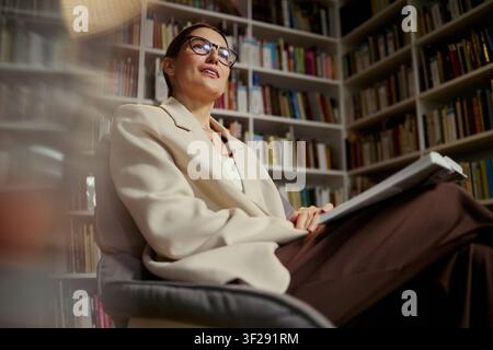 Caucasian middle aged woman sitting with open book in library, speaking during consultation or coaching session. Useful for education, mentoring, therapy, expertise marketing Stock Photo