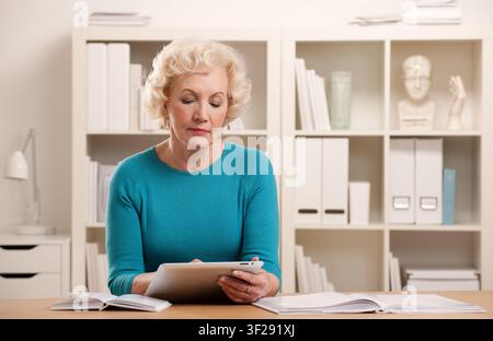 Senior female sitting at desk working on tablet in white office Stock Photo