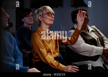 Caucasian middle aged businesswoman raising hand while attending seminar with diverse colleagues, signaling engagement and teamwork. Ideal for corporate training, leadership, workshop marketing Stock Photo