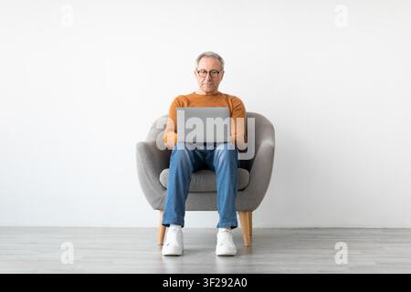Elderly Caucasian Man Using Laptop While Sitting On Comfortable Chair, Home Office Setting Stock Photo