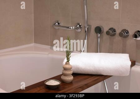 Rolled white towel on a wooden bath tray with spa essentials in a modern hotel bathroom, creating a relaxing wellness atmosphere Stock Photo