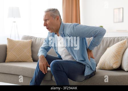 Elderly Caucasian Man Experiencing Back Pain While Sitting On Couch, Healthcare Concept Stock Photo
