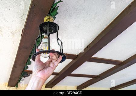 A man replaces a burned-out LED light bulb in a ceiling light fixture on the porch of a garden house. The image shows the process of unscrewing the ol Stock Photo