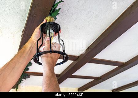 A man replaces a burned-out LED light bulb in a ceiling light fixture on the porch of a garden house. The image shows the process of unscrewing the ol Stock Photo