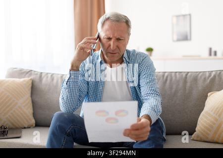 Focused Elderly Caucasian Man Analyzing Document While Talking On Phone, Home Office Stock Photo