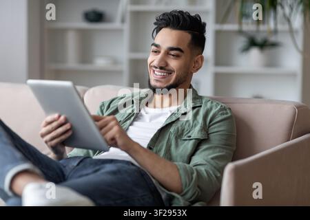 Smiling Young Man Using Tablet While Relaxing At Home, Living Room Interior Stock Photo