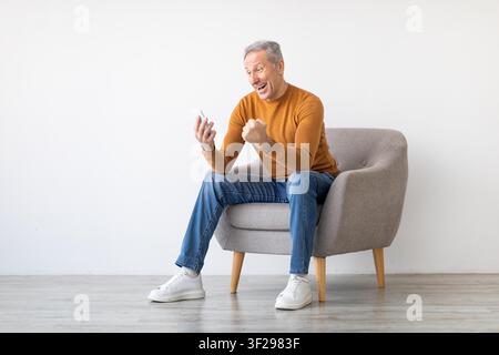 Happy Elderly Caucasian Man Celebrating Positive News While Using Smartphone, Home Setting Stock Photo