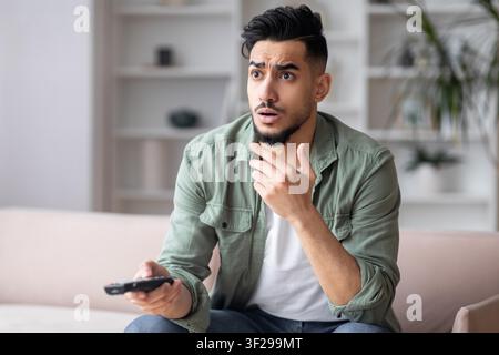 Worried Young Man With Beard Holding Remote Control Watching Tv At Home, Relaxed Concept Stock Photo