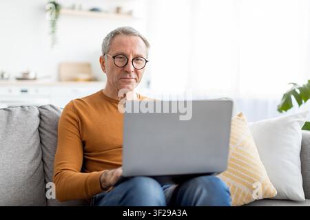 Senior Caucasian Man Browsing Laptop At Home, Focused, Modern Interior Stock Photo