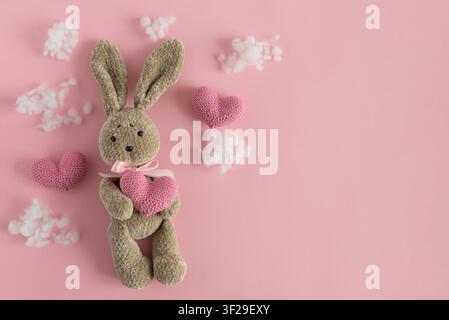Stuffed bunny toy holding a pink crocheted heart, surrounded by fluffy white clouds and additional crocheted hearts, all presented on a gentle pink ba Stock Photo