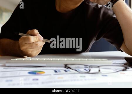 Close-up of a person thinking with a pen at an office desk featuring financial reports, keyboard, and copy space for business concepts. Stock Photo