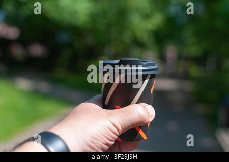 A man's hand holds a black takeaway paper coffee cup with a black lid while walking outside in a sunny green park Stock Photo