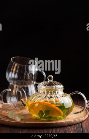 Glass teapot with warm herbal tea, fresh orange slice, and mint leaves, served on a wooden tray with two stacked empty glass cups and rock sugar Stock Photo