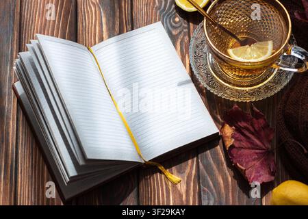 Opened notebook with blank lined pages and a golden bookmark lying next to a cup of hot lemon tea and a fall leaf on a dark rustic wooden table, illum Stock Photo