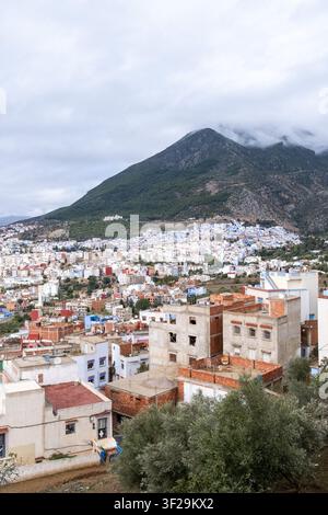 Elevated panoramic view of Chefchaouen cityscape, blue painted medina, dense rooftops and a large Rif mountain partly covered by low clouds in Chefcha Stock Photo