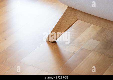 A detailed close up shot focusing on the angled oak wood leg and frame of a modern fabric sofa resting on a bright, polished natural hardwood parquet Stock Photo