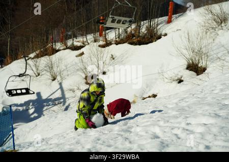 Child in a bright green snowsuit and colorful gloves rolls a large snowball while a small dog in a red coat frolics beside them on a snowy hillside wi Stock Photo