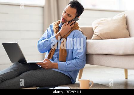 Man Using Laptop And Talking On Phone While Sitting On Floor, Home Office Environment Stock Photo