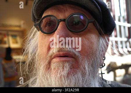 An senior man is shown with a beard, wearing round black glasses and a dark cap. His expression reflects varied emotions. A cross earring is visible o Stock Photo