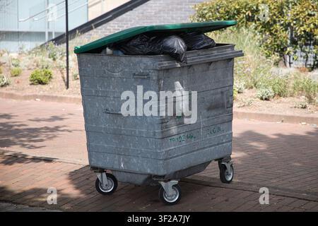 A large industrial wheelie bin with a green lid overflowing with black garbage bags on a paved walkway at Salford Quays, Salford, Manchester, UK. Stock Photo