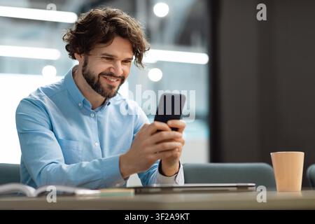 Smiling Caucasian Man Using Smartphone At Modern Office, Business Concept Stock Photo