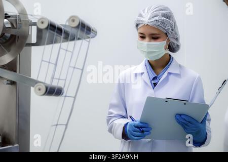 Asian woman scientist wearing sterile uniform checking pharmaceutical production line in factory. Concept of hygiene management and modern automatic p Stock Photo