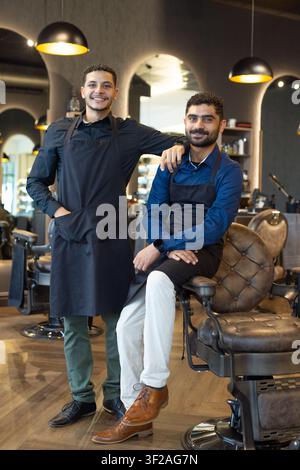 Two male barbers posing inside modern barbershop, wearing black aprons near tufted leather chairs Stock Photo