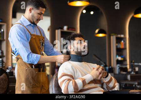 African American male barber securing neck strip, male client sitting in chair at shop, copy space Stock Photo