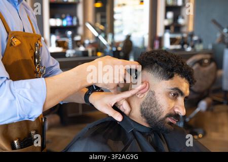 Male barber trimming male client's hair at shop using electric clippers, scissors in leather apron Stock Photo