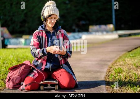 Woman sitting on skateboard, wearing headphones and looking at smartphone in a skate park Stock Photo