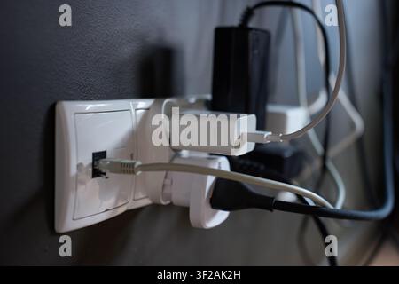 Multiple chargers and cables connected to wall socket behind electronic equipment. Concept of electrical connections and cable organization Stock Photo