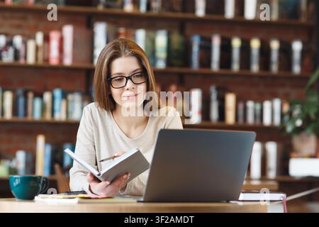 Focused Young Woman Writing In Notebook While Using Laptop At Cafe, Business Concept Stock Photo