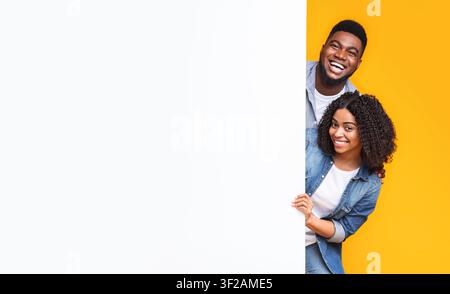 Happy Black And White Young Couple Smiling And Peeking From Behind White Board, Yellow Background Stock Photo