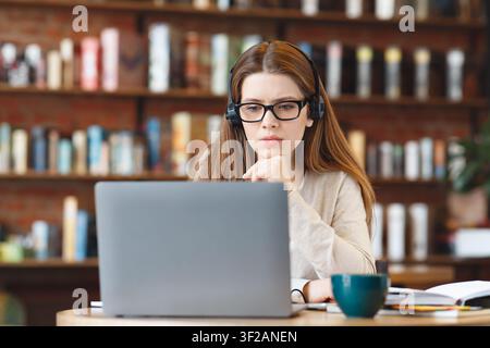 Focused Young White Woman Working On Laptop With Headphones In Modern Coffee Shop Setting Stock Photo