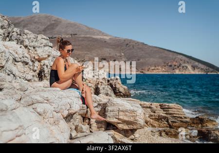 Young woman in black swimsuit sitting on rocky coast of Cephalonia Greece reading e-book, happy female traveler relaxing by blue Ionian Sea with sceni Stock Photo
