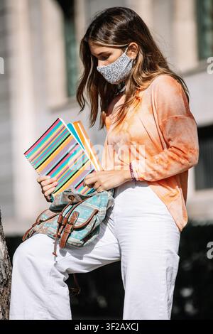 Young female in fabric mask putting planners inside bag on blurred background of university building on summer day in Madrid, Spain Stock Photo