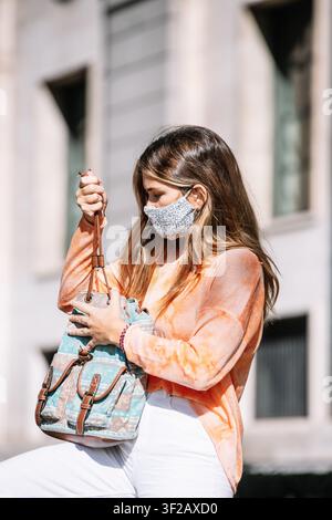 Young female in fabric mask putting planners inside bag on blurred background of university building on summer day in Madrid, Spain Stock Photo